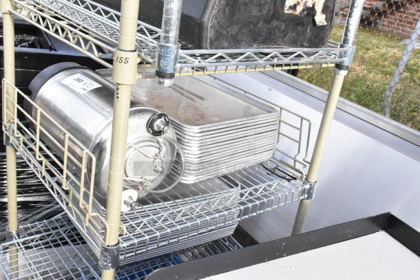 Wire shelving unit on commercial casters holding metal trays and a steel container. Includes trash can. Buyer disassembly required.