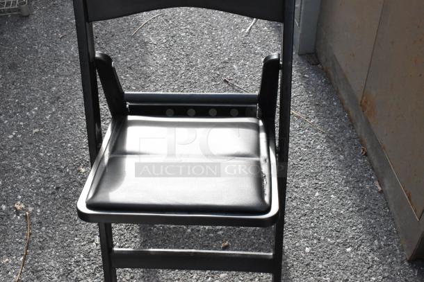 Black folding chair on asphalt, with a padded seat. Part of a lot of 56 white and black chairs in good condition.