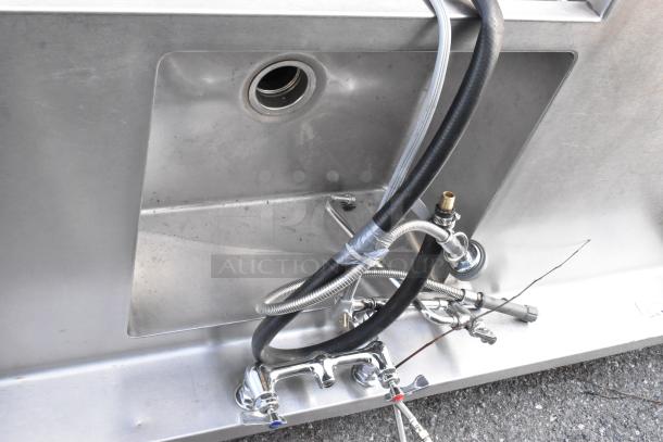Stainless steel single bay L-shaped sink with faucets and plumbing attachments, showing light wear.