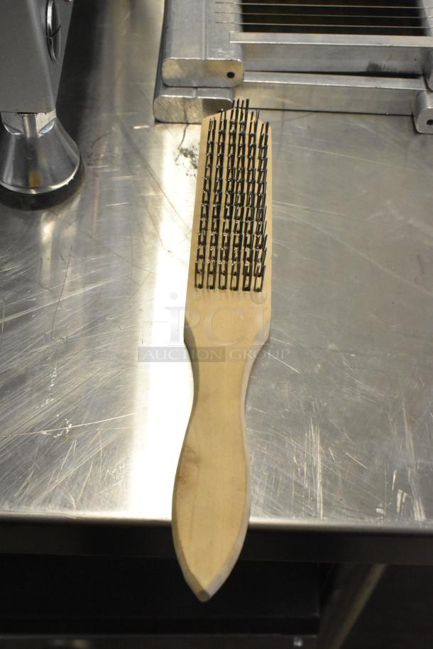 Wooden handle coarse cleaning brush with sturdy metal bristles, in good condition, on a metallic surface.