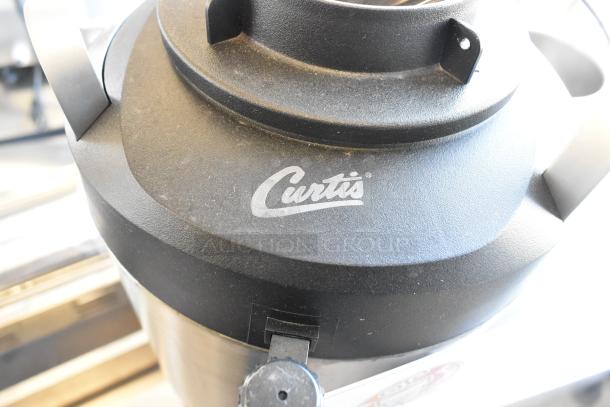 Stainless steel Curtis beverage dispenser, top view, featuring brand logo. Black lid, sturdy handles, excellent condition.