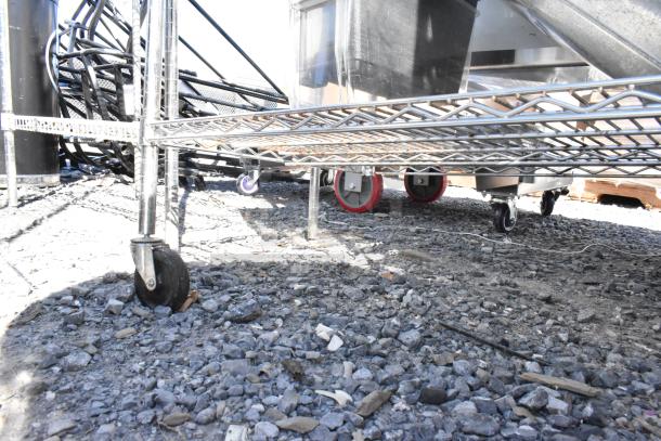 Chrome finish 4-tier wire shelving unit with contents on commercial casters, situated outdoors on gravel. Buyer must dismantle.