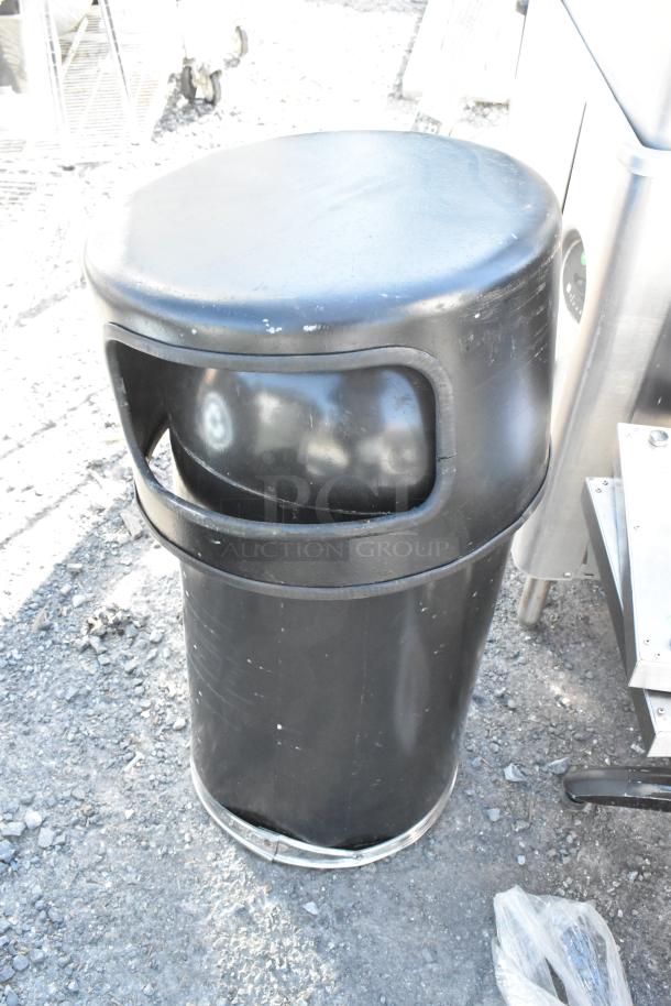 Black cylindrical trash can shell, outdoor condition, visible surface wear, sturdy plastic construction.