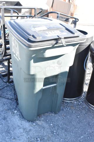 Green 65-gallon trash can with contents, marked with "U1 354228" and "TRASH" label, outdoor condition.