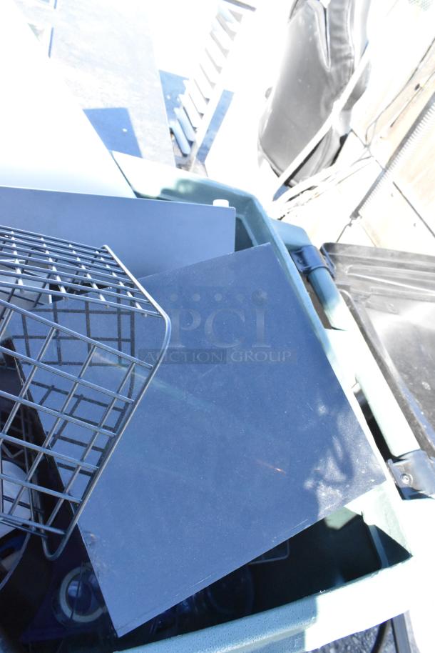 Green poly trash can filled with various black items, including a metal grid. Lid partially open.