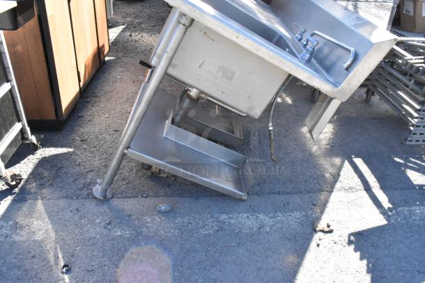 Stainless steel single bay sink with counter, backsplash, and under shelf; slightly tilted, visible pipe and faucet.