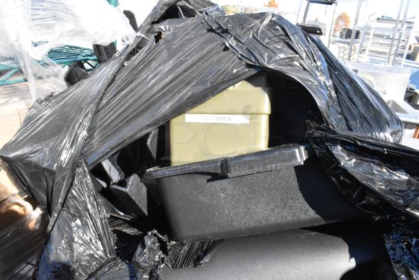 Pallet lot with various items including a trash can and bus bin, wrapped in black plastic. "Cold Brew" label visible. Mixed condition.