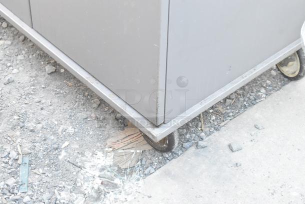 Gray 2 Door Cart with commercial casters, minor wear visible, positioned on gravel and concrete surface.