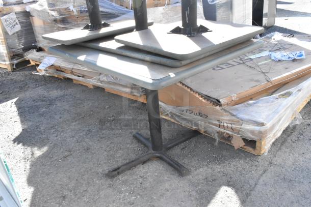 Four metal tables with square tops and black pedestal bases, stacked outdoors on a pallet. Slight wear visible.