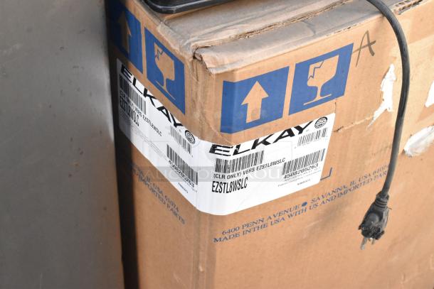 Cardboard box with Elkay label, showing part number EZSTL8WSLC, next to a metal surface. Box corners slightly worn. Plug cable visible.