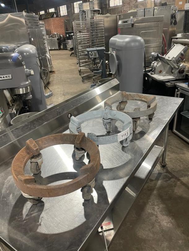 Three heavy-duty round trash can dollies on casters in a commercial kitchen setting. Visible wear and rust.