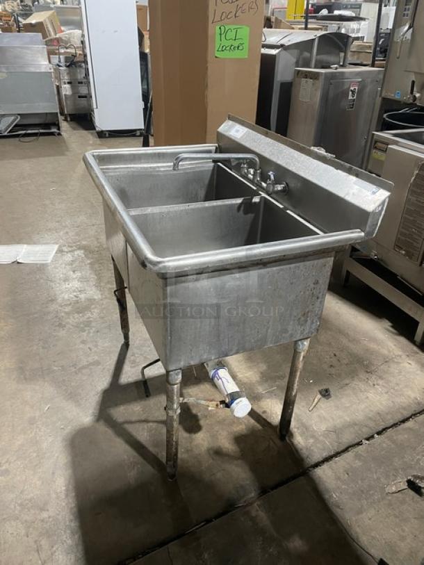 All stainless steel two-compartment dishwashing/prep sink with faucet and legs, showing signs of use.
