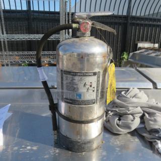 Stainless steel One K Guard fire extinguisher with hose, visible instructions and inspection tag, in used condition.