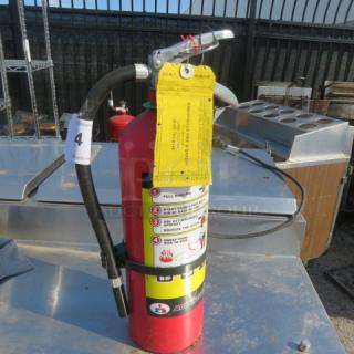 Red ABC fire extinguisher, with a hose and metal handle, safety instructions visible. Yellow inspection tag attached.