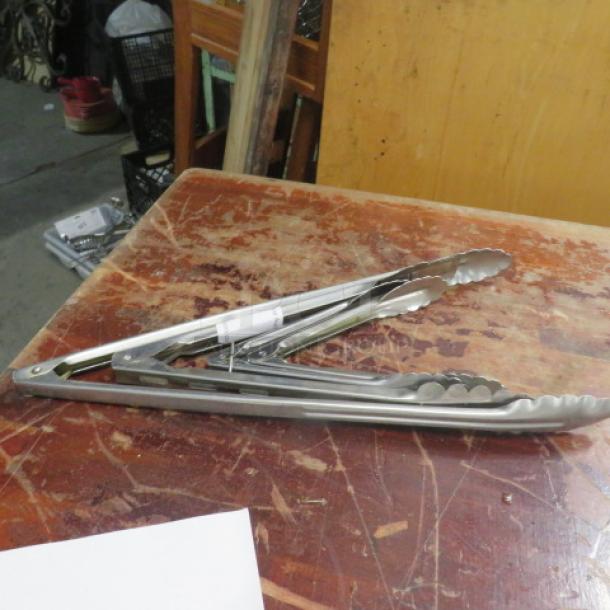 Assorted stainless steel tongs on a wooden table, slightly used, with a paper tag indicating "4XBID" attached.