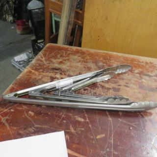 Assorted stainless steel tongs on a wooden table, slightly used, with a paper tag indicating "4XBID" attached.