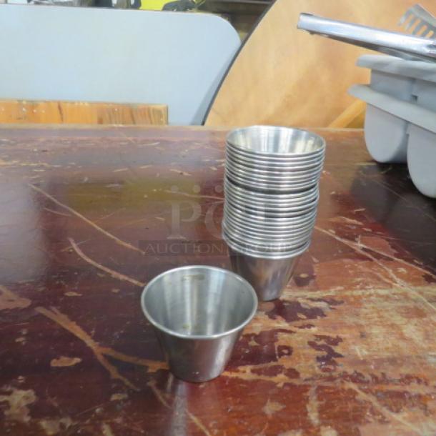 Lot of 25 stainless steel condiment cups, stacked on a wooden surface. Cups appear in good condition, showing minor wear.