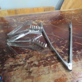 Assorted kitchen tongs on a wooden table, includes multiple styles with varying lengths and gripping ends, in used condition.