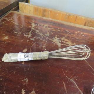 Stainless steel whip, wrapped in plastic, resting on a worn wooden table. Attached label with illegible text.