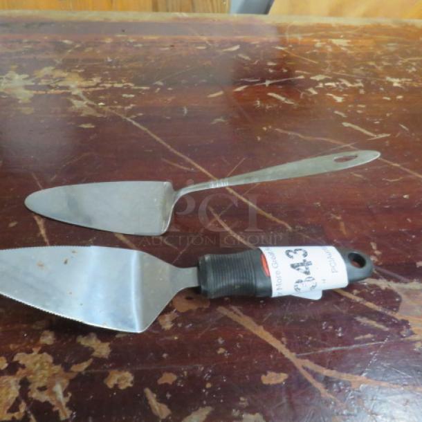 Two assorted pizza/pie servers with stainless steel blades and black handles on a wooden table. One has a tag labeled "143."