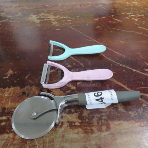 Assorted kitchen utensils including a pizza cutter and two peelers, one pink and one blue. Item tag reads "346."