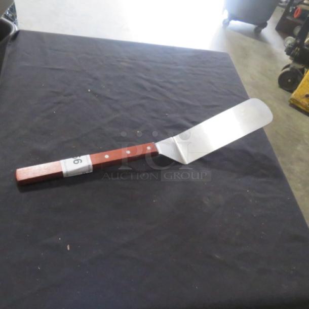 A stainless steel hamburger turner with a wooden handle, in good condition, displayed on a dark fabric surface.