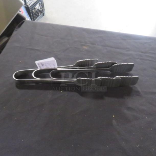 Assorted stainless steel tongs on table, featuring ridged grips and a labeled tag, indicating "3XBID."