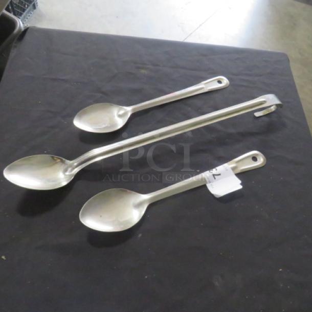 Assorted stainless steel cooking spoons, three pieces, various sizes, with hanging holes. Good condition, auction tag attached.