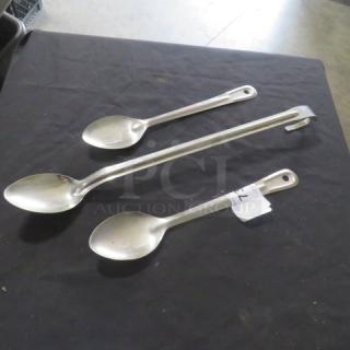 Assorted stainless steel cooking spoons, three pieces, various sizes, with hanging holes. Good condition, auction tag attached.