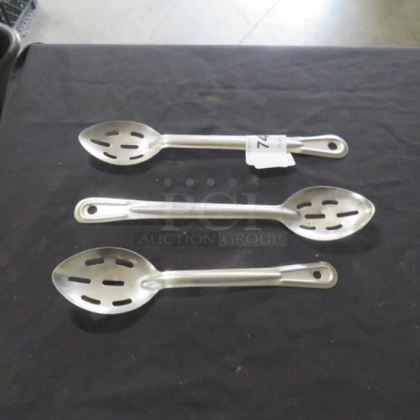 Assorted slotted spoons on black surface; three sizes, metal construction, slightly used condition, auction lot tag visible.