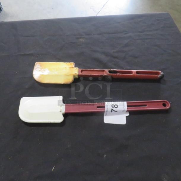 Two assorted spatulas on a table; one has a yellow-stained head, and the other is white. Both have red handles. Auction tag marked "78."