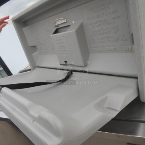 Gamco wall mount baby changing station, grey, open position, safety strap visible, installation and safety instructions embossed on interior.