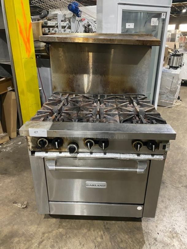 Garland commercial stainless steel gas-powered 6-burner range/oven combo with backsplash, overhead salamander shelf, and wire oven rack on casters.