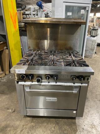 Garland commercial stainless steel gas-powered 6-burner range/oven combo with backsplash, overhead salamander shelf, and wire oven rack on casters.
