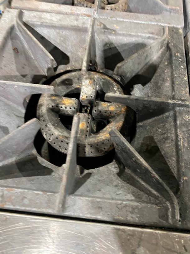 CPG Commercial Stainless Steel 4-burner range detail, showing gas burners. Features backsplash, salamander shelf, and casters. Signs of use visible.