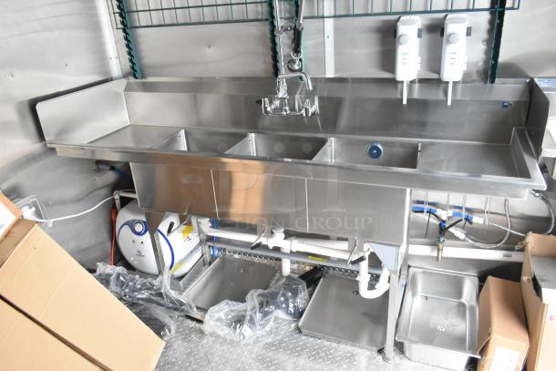 Stainless steel triple sink setup in a fully outfitted food trailer, featuring soap dispensers and plumbing fixtures.