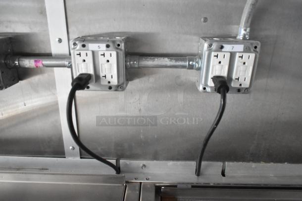 Electrical outlets inside a food trailer, labeled "5" and "7," with cords plugged in, on a metal wall surface.