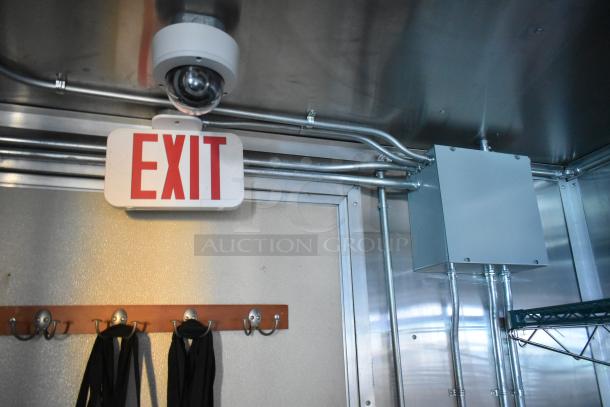 Food trailer interior showing a mounted EXIT sign, security camera, and metal shelving. Visible wiring and hooks beneath.
