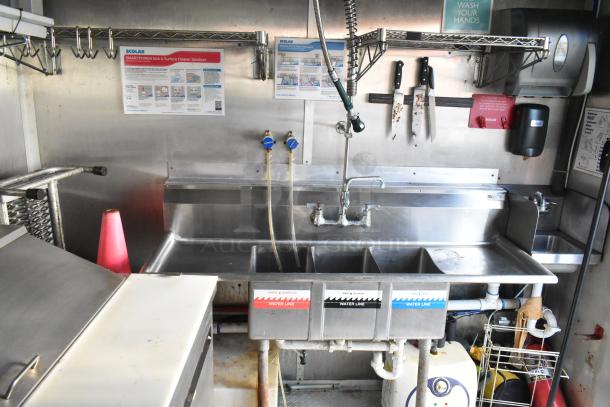 Stainless steel sink and prep area in a brand new food trailer, featuring labeled water lines, knives, sanitizer info, and equipment.