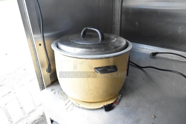 Electric rice cooker with lid and power cord on a metal countertop inside a food trailer kitchen.