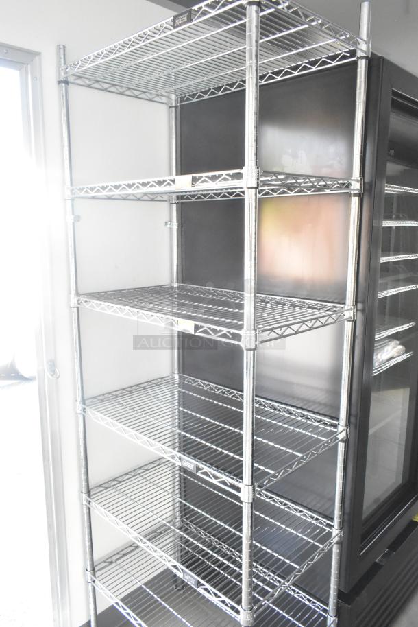Wire shelving unit in food trailer, stainless steel, adjacent to black reach-in cooler. Ideal for commercial storage.