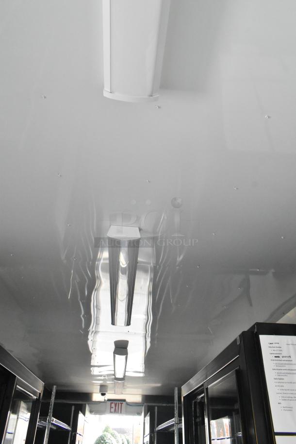 Interior view of a fully equipped unbranded food trailer with lighting, wire shelving, and commercial-grade appliances. Exit sign visible.