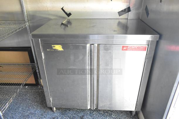 Stainless steel cabinet with double doors and Atlantic Coast sticker, next to wire shelving inside a mobile kitchen.