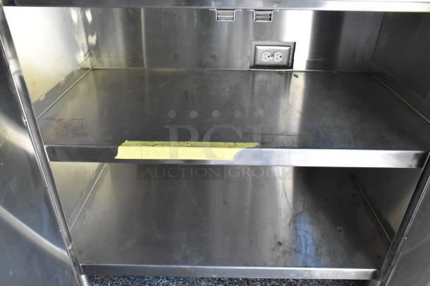 Stainless steel interior of food trailer with power outlet and two shelves. Yellow tape labeled "Dead Batteries Only."