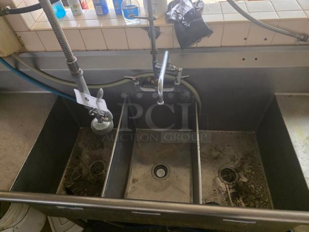 Stainless steel 3 compartment sink with industrial faucet, visibly worn condition, features attachment hoses and mounted on a tiled backsplash.