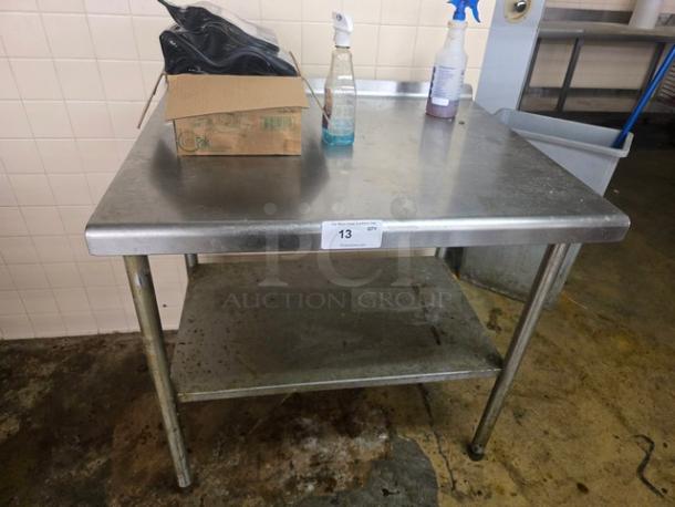 Stainless steel work table with a lower shelf, showing wear. Includes cleaning supplies on top. Auction tag indicates item number 13.