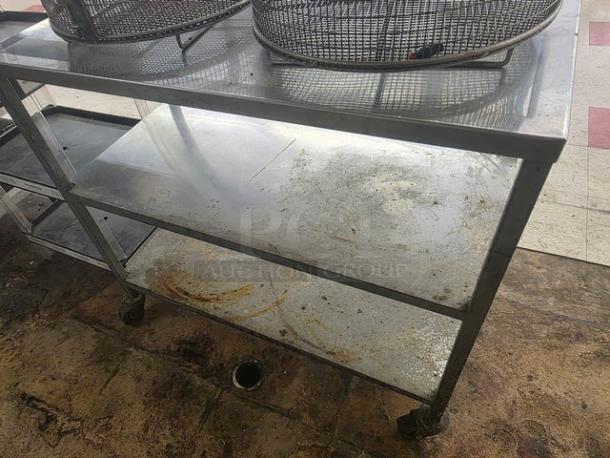 Stainless steel cart with three shelves, visible wear. Contents on top not included. Sturdy design with casters for mobility.