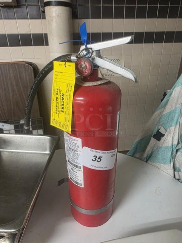 Red fire extinguisher with metal handle, yellow inspection tag, and gauge. Appears in good condition.