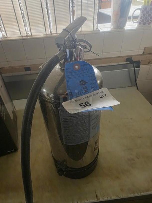 Industrial fire extinguisher, silver metal body, with attached hose. Features inspection tag and auction label marked "56".