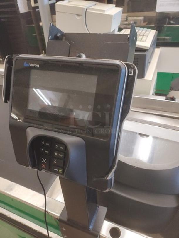 Supermarket checkout counter with Verifone card reader, metal finish, slight wear. Ideal for retail use.